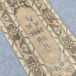 May include: A vintage runner rug with a central beige field decorated with floral patterns, framed by brown and cream stripes. The rug incorporates pink and green details, offering a warm, welcoming appearance. The design suggests a traditional, handcrafted style.
