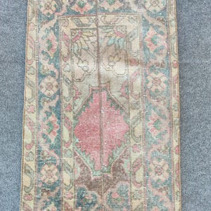 Op de afbeelding: Een rechthoekig, versleten tapijt met een vervaagde, vintage uitstraling. Het tapijt heeft een centrale ruitvorm in roze, omgeven door bloemen en geometrische patronen in tinten blauw, groen en beige. De randen van het tapijt zijn gerafeld.
