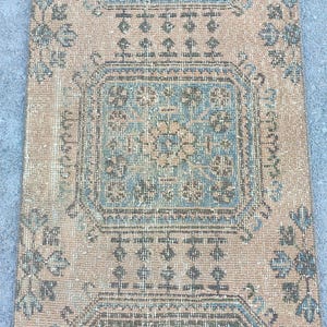 May include: A rectangular, vintage-style rug with a distressed appearance. The rug features a central design in shades of blue and beige, surrounded by a border with floral and geometric patterns. The rug's color palette includes muted tones of beige, blue, and brown.