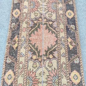 May include: A long, narrow runner rug featuring a central geometric pattern in shades of dusty pink, olive green, and light blue on a beige background, framed by dark charcoal borders with repeating floral and diamond motifs throughout the design.
