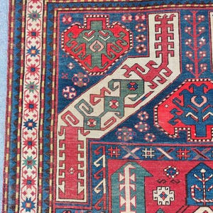 May include: Detail of a traditional rug with a rich, intricate design. The rug's main field is a deep blue, contrasted by red, green, and cream-colored geometric patterns. The border showcases a repeating floral pattern.