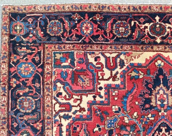 May include: Close-up of a section of a Persian rug with a detailed floral pattern. The rug features a cream-colored central field with red, blue, and pink geometric designs. A dark blue border with floral motifs frames the rug's edge.