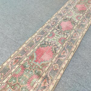 May include: A long, rectangular rug with a floral pattern in shades of pink, green, and beige. The rug has a distressed, vintage look, with a central design and borders. The rug's dimensions are not visible in the image.