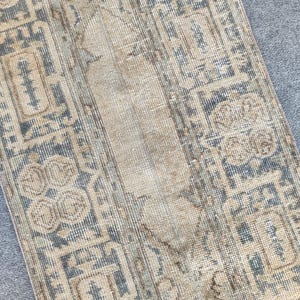 May include: A rectangular, distressed rug with a faded, vintage aesthetic. The rug features a central beige field with a repeating pattern of muted blue and beige geometric shapes and floral motifs. The rug's edges show signs of wear.