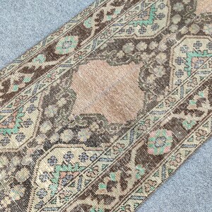 Könnte beinhalten: Ein antiker Läuferteppich mit einem abgenutzten, Vintage-Aussehen. Der Teppich hat ein zentrales pfirsichfarbenes Feld, das von einem Rand aus braunen, beigen und türkisfarbenen Blumenmustern umgeben ist. Das Design des Teppichs ist symmetrisch und detailliert.