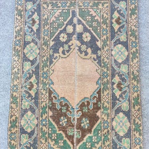 May include: A rectangular woven rug featuring a central peach-coloured medallion surrounded by dark blue, green, and brown geometric floral patterns. The border displays intricate repeating motifs in muted tones with a fringed edge at the top.