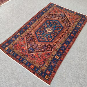 May include: A rectangular, hand-knotted rug with a central geometric medallion in shades of blue, brown, and red. The rug features intricate floral and geometric patterns, with a dark blue border and a cream fringe.