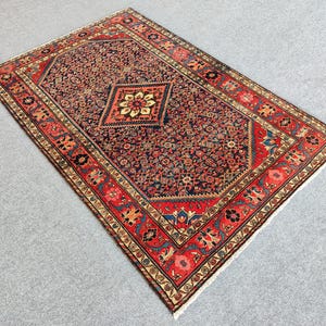 Peut inclure: Un tapis rectangulaire pr&eacute;sentant un champ central bleu marine fonc&eacute; rempli de motifs floraux complexes et un m&eacute;daillon central en losange contenant une fleur cr&egrave;me et jaune. La bordure est d'un rouge vif avec des motifs floraux.