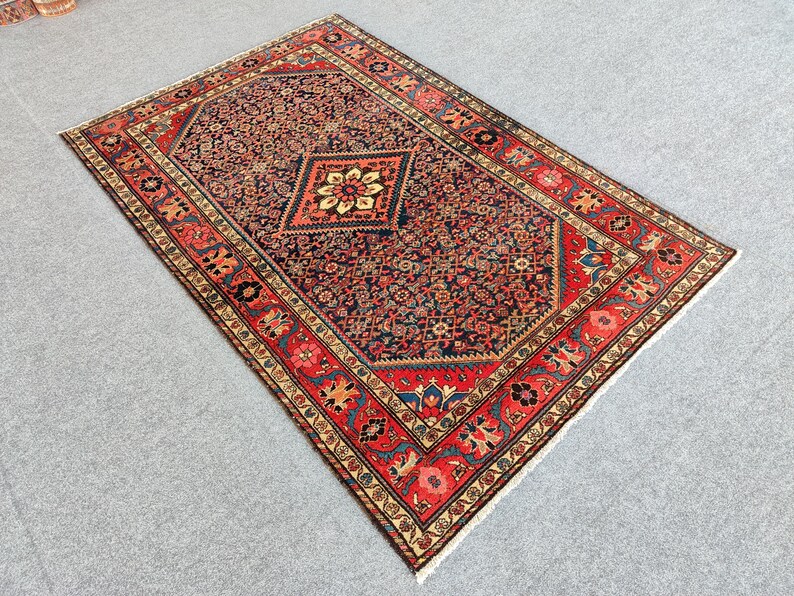 Peut inclure: Un tapis rectangulaire pr&eacute;sentant un champ central bleu marine fonc&eacute; rempli de motifs floraux complexes et un m&eacute;daillon central en losange contenant une fleur cr&egrave;me et jaune. La bordure est d'un rouge vif avec des motifs floraux.