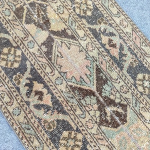 May include: Close-up of a vintage rug with a distressed appearance. The rug features a repeating pattern of floral and geometric designs in muted tones of beige, brown, navy, and pink. The rug's texture is visible, suggesting a woven construction.