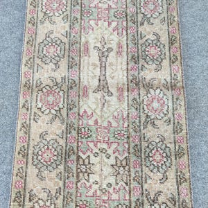 Pode incluir: Um tapete retangular com fundo bege e um padrão floral repetido em rosa, verde e castanho. O tapete tem um design central com um motivo vertical estilizado e bordas com elementos florais e geométricos.