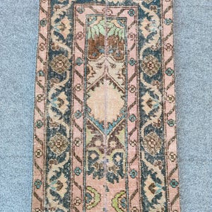 May include: A rectangular, vintage rug with a faded, distressed look. The rug has a central design in pink, brown, and green, framed by a dark teal border with floral patterns. The design is symmetrical. The rug is likely made of wool.