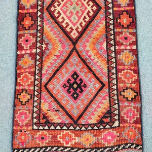 May include: A rectangular, hand-woven rug with a geometric design. The rug features a central diamond pattern in shades of orange, pink, and black, framed by a border of repeating floral motifs in red and orange. The rug's dimensions are not visible.