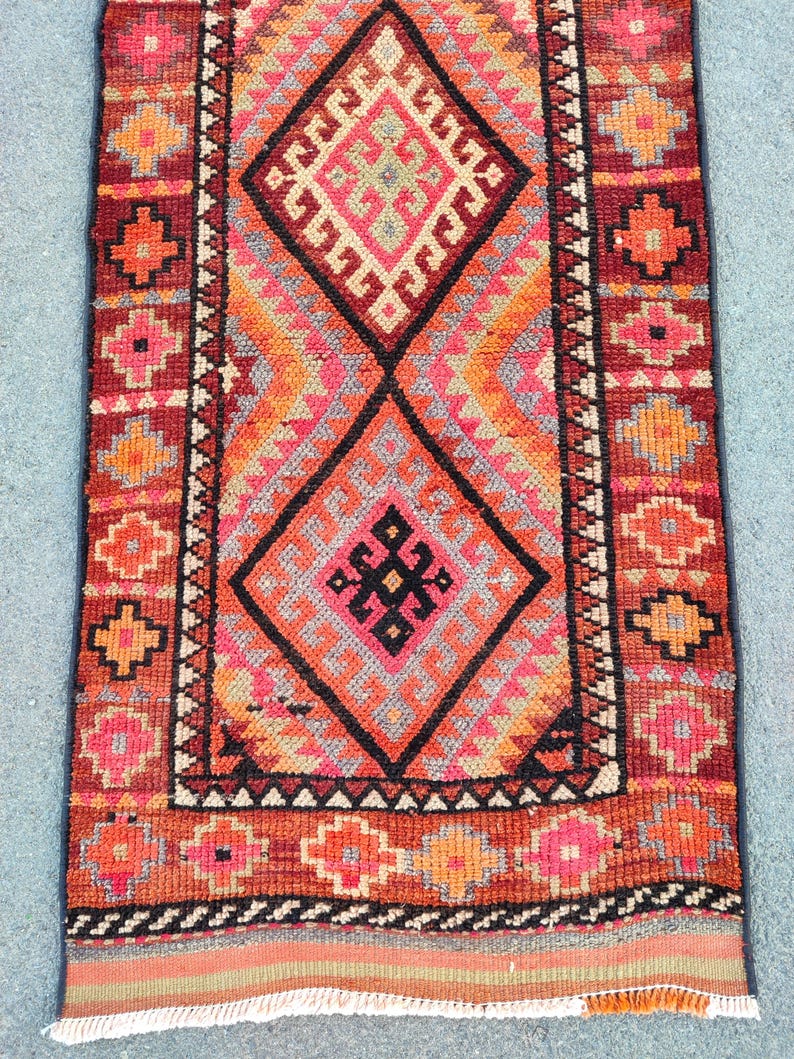 May include: A rectangular, hand-woven rug with a geometric design. The rug features a central diamond pattern in shades of orange, pink, and black, framed by a border of repeating floral motifs in red and orange. The rug's dimensions are not visible.