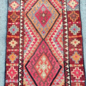 May include: A rectangular, hand-woven rug with a geometric pattern. The rug features a central diamond design in shades of red, orange, and gray, framed by a dark red border with floral motifs. The rug's dimensions are not visible.