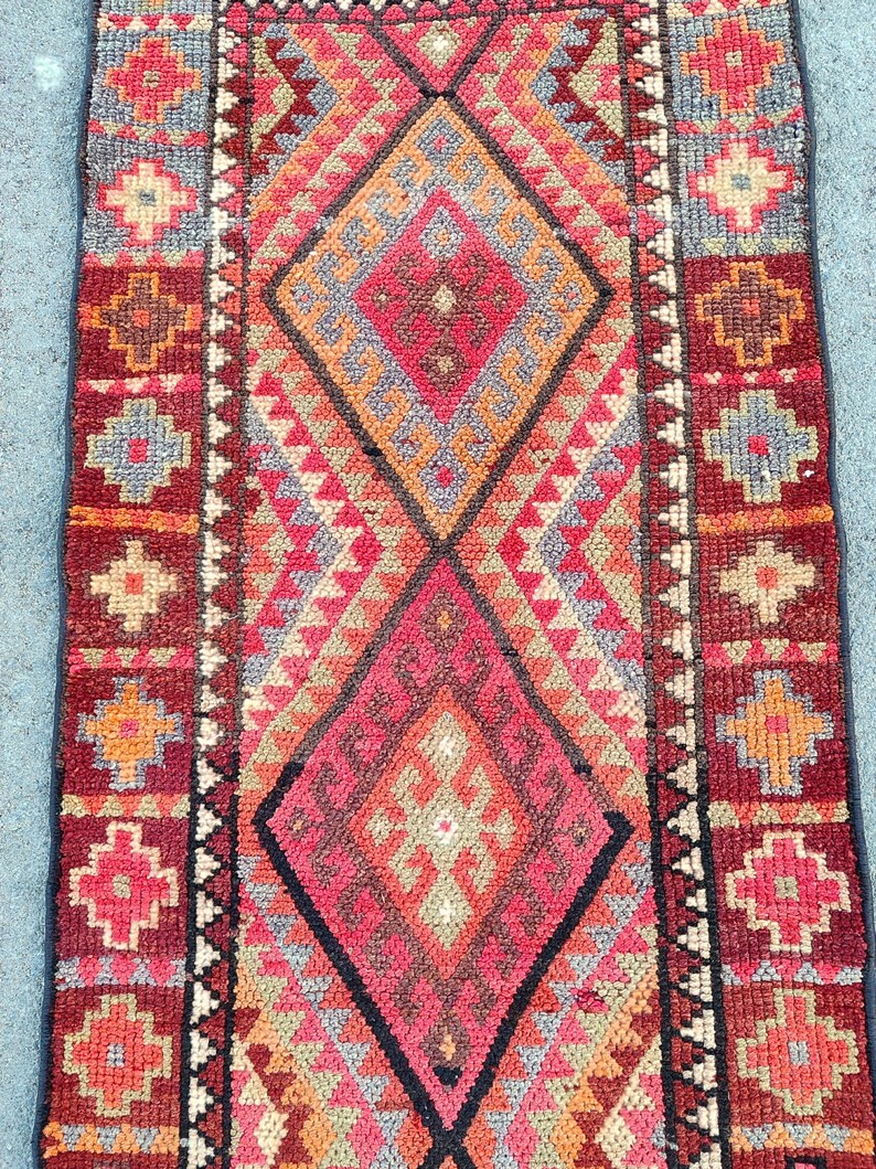 May include: A rectangular, hand-woven rug with a geometric pattern. The rug features a central diamond design in shades of red, orange, and gray, framed by a dark red border with floral motifs. The rug's dimensions are not visible.