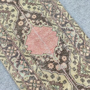 May include: A vintage rug with a worn, faded look. It has a central peach-coloured section, framed by olive green, brown, and floral patterns. The rug's design is symmetrical and detailed, with a classic aesthetic.
