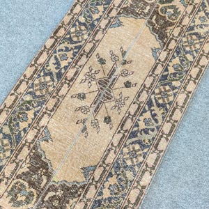 Può includere: Un tappeto rettangolare in stile vintage con un campo centrale beige e intricati motivi floreali e geometrici. I bordi presentano accenti marrone scuro e blu, creando un aspetto classico e invecchiato. Il tappeto è probabilmente realizzato in materiale tessuto.