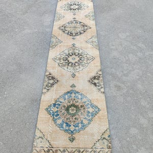 May include: A long, rectangular rug with a beige background and intricate geometric patterns in shades of blue, green, and black. The rug features a central diamond pattern and borders with floral motifs, suitable for a hallway or entryway.