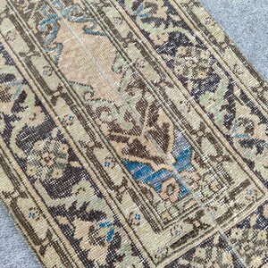 May include: Close-up of a section of a vintage rug with a distressed, faded appearance. The rug features a central beige field with floral and geometric patterns in shades of brown, blue, and green. The edges and borders are darker, with a similar pattern.