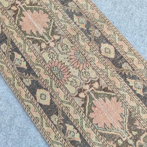 May include: A rectangular rug with an intricate floral pattern. The rug features a color palette of muted pink, beige, and dark brown. The design includes stylized flowers and geometric shapes, creating a vintage aesthetic. The rug is likely made of woven material.