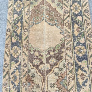 May include: A rectangular runner rug featuring a central beige medallion surrounded by brown, dark blue, and green floral patterns. The border displays geometric motifs in dark blue and tan, with a weathered texture throughout the woven surface.
