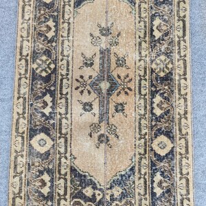 May include: A rectangular, vintage rug with a central beige panel and detailed dark blue and brown borders. The rug displays floral and geometric patterns, giving it a distressed, antique look. The rug's dimensions are not visible.