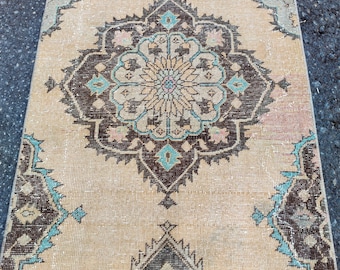 May include: A rectangular beige rug with a central medallion design. The rug has a symmetrical pattern with brown, teal, and pink accents. The border features a darker brown and teal design. The rug is likely woven.