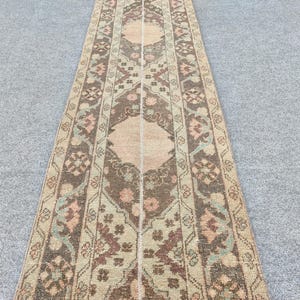 May include: A rectangular, vintage-style runner rug with a central design in shades of brown, beige, and peach. The rug features floral and geometric patterns, with a border in complementary colours. The rug is placed on a grey surface.