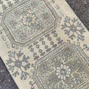 Puede incluir: Una alfombra rectangular con un patrón geométrico repetitivo en tonos beige, gris y azul. El diseño presenta motivos florales y geométricos, creando una estética vintage o antigua. La textura de la alfombra parece suave y acogedora.