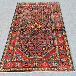 Peut inclure: Un tapis rectangulaire pr&eacute;sentant un champ central bleu fonc&eacute; avec un motif floral r&eacute;p&eacute;titif et un m&eacute;daillon central en losange. Le tapis est encadr&eacute; par une large bordure rouge orn&eacute;e de motifs floraux complexes et de d&eacute;tails cr&egrave;me.
