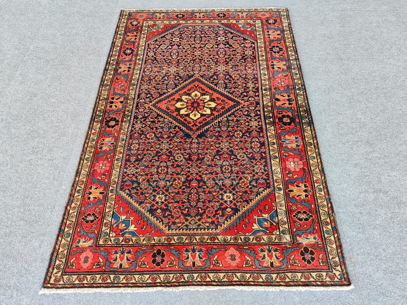 Peut inclure: Un tapis rectangulaire pr&eacute;sentant un champ central bleu fonc&eacute; avec un motif floral r&eacute;p&eacute;titif et un m&eacute;daillon central en losange. Le tapis est encadr&eacute; par une large bordure rouge orn&eacute;e de motifs floraux complexes et de d&eacute;tails cr&egrave;me.
