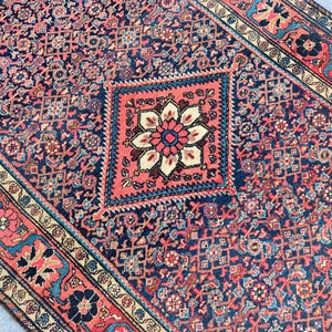 Peut inclure: Un tapis rectangulaire pr&eacute;sentant un champ bleu fonc&eacute; avec un motif floral g&eacute;om&eacute;trique dense et r&eacute;p&eacute;titif. Un m&eacute;daillon central en forme de losange rose corail contient une grande fleur &agrave; p&eacute;tales multiples. La bordure affiche des motifs floraux.