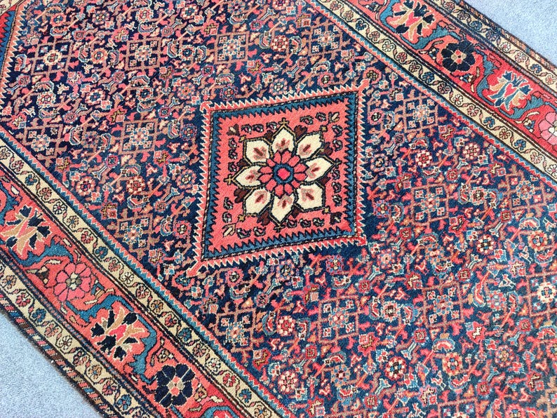 Peut inclure: Un tapis rectangulaire pr&eacute;sentant un champ bleu fonc&eacute; avec un motif floral g&eacute;om&eacute;trique dense et r&eacute;p&eacute;titif. Un m&eacute;daillon central en forme de losange rose corail contient une grande fleur &agrave; p&eacute;tales multiples. La bordure affiche des motifs floraux.