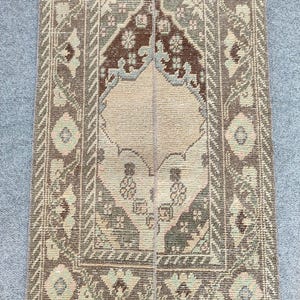 Peut inclure: Un tapis rectangulaire de type chemin de table avec un médaillon central beige entouré d'un champ brun foncé. La bordure présente des motifs géométriques dans des tons sourds de vert sauge, bleu clair et crème, avec une couture verticale visible.