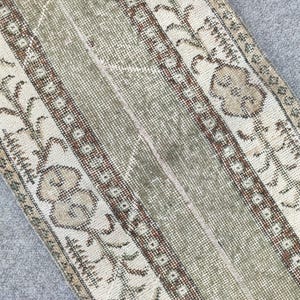 May include: Close-up of a distressed, rectangular rug with a faded, vintage aesthetic. The rug features vertical stripes in shades of beige, olive green, and brown, with floral and geometric patterns. The rug's design suggests a traditional, handcrafted style.