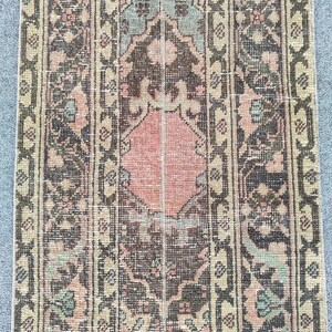 May include: A rectangular, vintage-style rug with a central pink diamond shape and intricate floral patterns in shades of brown, beige, and green. The rug has a distressed look, with faded colors and a worn texture.