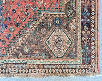 Op de afbeelding: Een geweven tapijt met een levendig koraalrood centraal veld met ingewikkelde geometrische en florale motieven in blauw, groen en crème. Het tapijt heeft een donkerbruine rand met een ruitvormig medaillon en een franje aan de onderkant.