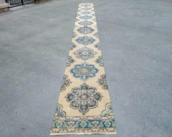 May include: A long, narrow beige runner rug with an intricate pattern of blue, brown, and green floral medallions. The rug has a border with a repeating geometric design. The rug is placed on a grey surface.