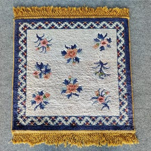 May include: Square rug with a cream centre featuring floral patterns in blue and coral. The rug has a dark blue border with a geometric design and a gold fringe.