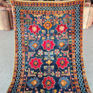 Op de afbeelding: Een rechthoekig vloerkleed met een diepblauwe achtergrond, voorzien van een symmetrisch patroon van grote rode bloemmotieven, kleinere groene en gele geometrische vormen en een brede rand met complexe blauwe en oranje zigzag- en ruitpatronen.