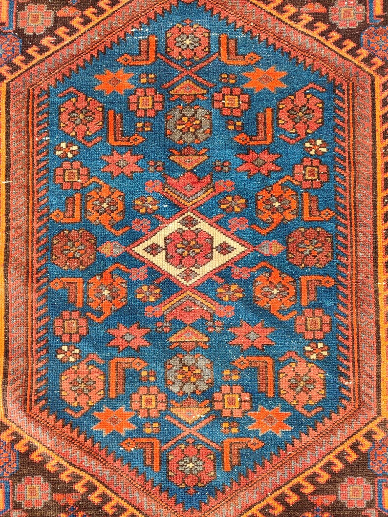 May include: A detailed, rectangular rug with a central blue field. The rug features a complex geometric pattern in shades of orange, red, and cream. The border is a dark brown with a repeating design. The rug's design is symmetrical.