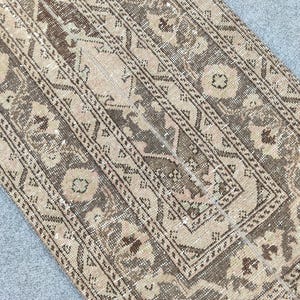 Könnte beinhalten: Ein antiker Teppich mit einem verblichenen, abgenutzten Aussehen. Der Teppich hat ein zentrales rechteckiges Design mit floralen und geometrischen Mustern in Braun-, Beige- und verblassten Rosatönen. Der Rand ist dunkelbraun.
