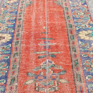 Könnte beinhalten: Ein abgenutzter, rechteckiger Teppich mit einem roten Mittelfeld und gemusterten Bordüren. Die Bordüren weisen blaue, grüne und beige Blumenmuster auf. Das Design des Teppichs umfasst geometrische und florale Motive und schafft eine Vintage-Ästhetik.