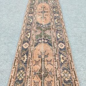 May include: A long, narrow, vintage rug with an intricate floral pattern. The rug features a central peach-colored field with a repeating floral design, bordered by navy blue and green accents. The rug's design is symmetrical and detailed.