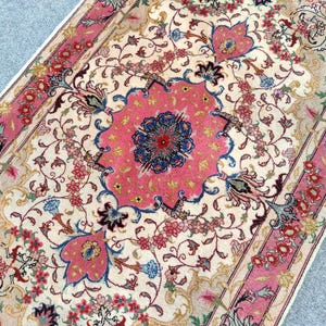 May include: A rectangular area rug featuring a cream-colored field with an intricate floral pattern in shades of pink, blue, and green. A large central pink medallion with a dark blue floral center is surrounded by scrolling vines and blossoms.