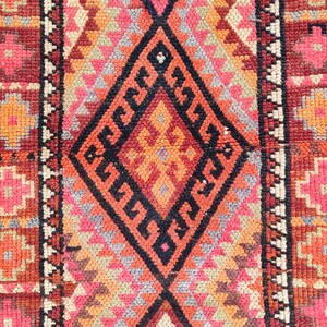 May include: A rectangular, hand-woven rug with a central diamond pattern in black, orange, and red. The rug features geometric designs in shades of pink, orange, and gray, with a dark red border. The rug's dimensions are not visible.