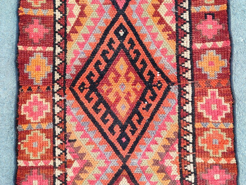 May include: A rectangular, hand-woven rug with a central diamond pattern in black, orange, and red. The rug features geometric designs in shades of pink, orange, and gray, with a dark red border. The rug's dimensions are not visible.