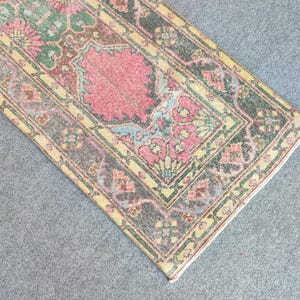 Pode incluir: Um tapete retangular em estilo vintage com um medalhão rosa central e padrões florais em verde, amarelo e azul. O tapete tem um aspeto envelhecido com uma borda cinza escuro e uma aparência desbotada.