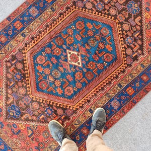 May include: A rectangular rug with a central blue geometric design, surrounded by a border of red, brown, and blue patterns. The rug features intricate floral and geometric motifs. The rug's dimensions are not visible in the image.